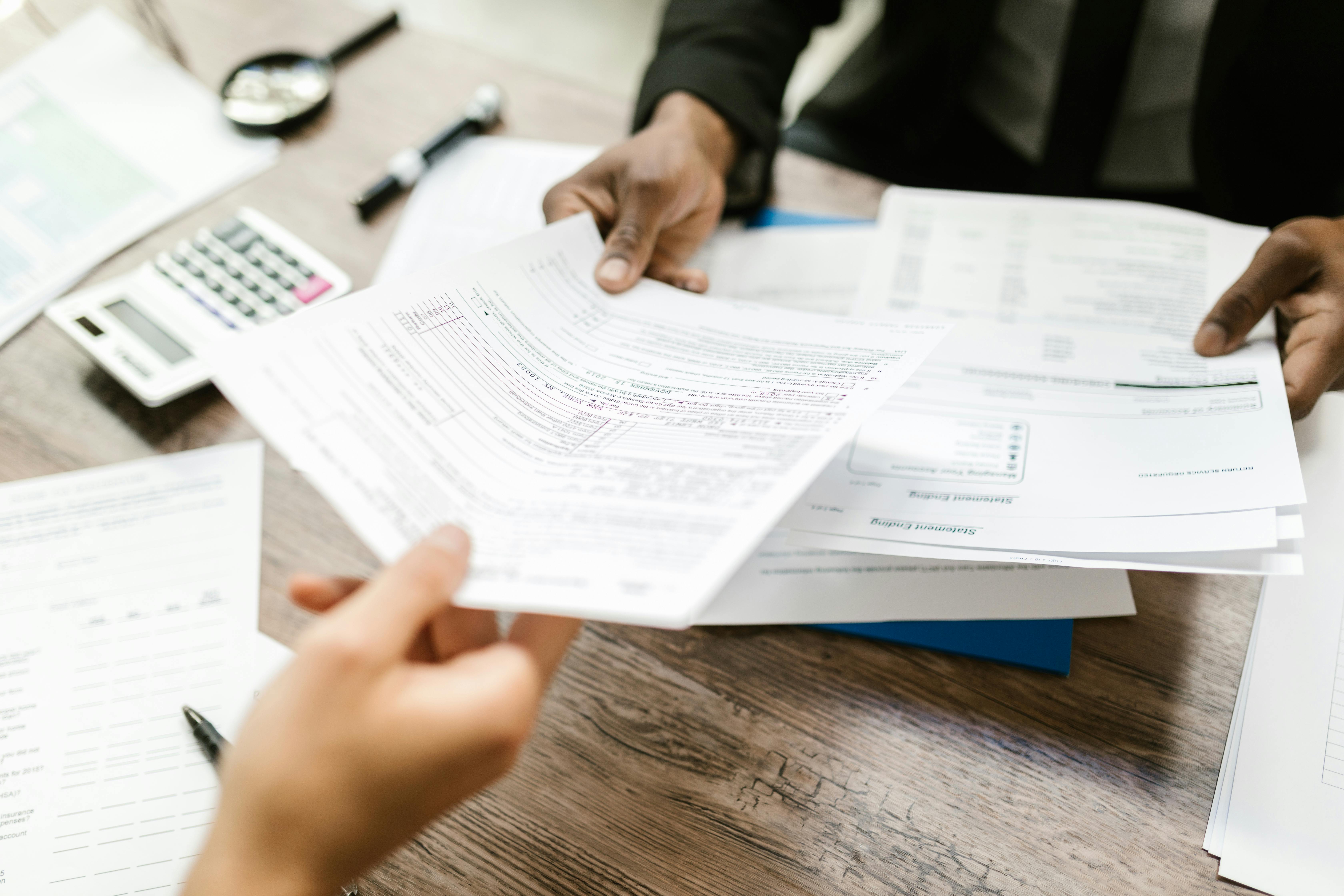 Close-up of business professionals reviewing and exchanging tax and payroll documents at a desk with paperwork, a calculator, and a pen.